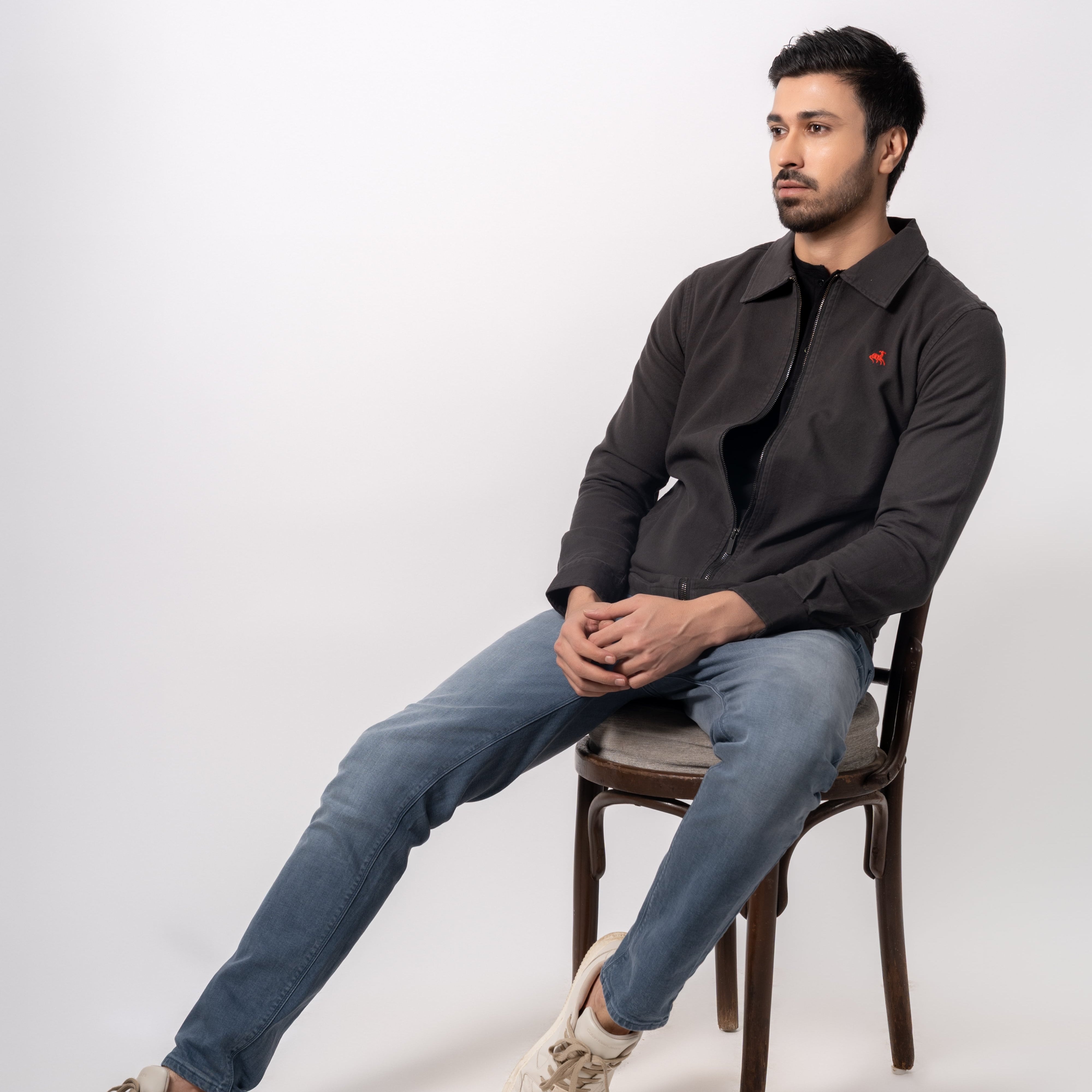 Man sitting on a chair wearing a dark jacket and jeans against a white background