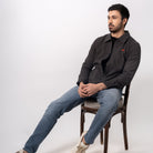 Man sitting on a chair wearing a dark jacket and jeans against a white background